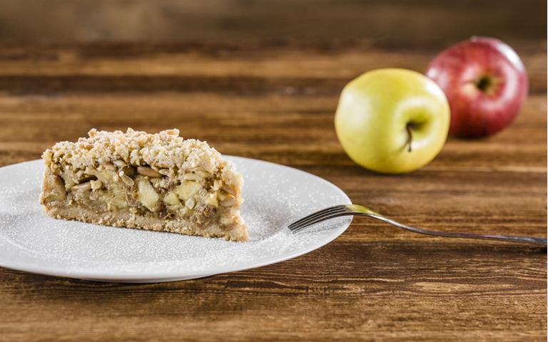 Una fetta di torta di mele con crumble su un piatto bianco spolverato di zucchero a velo; sullo sfondo due mele, una rossa e una gialla, appoggiate su un tavolo di legno.