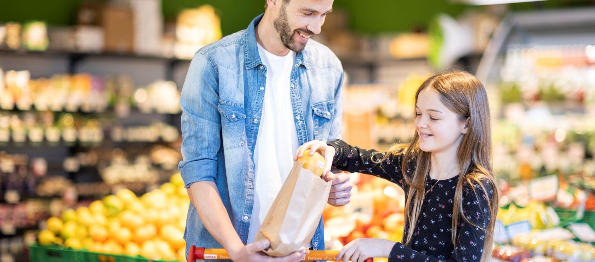 Un uomo e una bambina acquistano insieme delle mele in un supermercato e le mettono in un sacchetto di carta – sullo sfondo si vede frutta fresca.