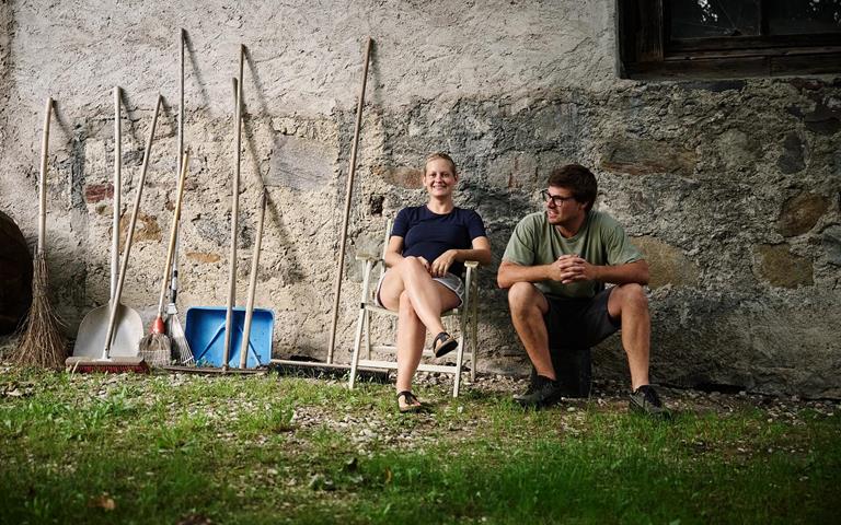 Andrea Erlacher e Jacob Gamper siedono insieme davanti al maso, accanto a diversi attrezzi da lavoro.
